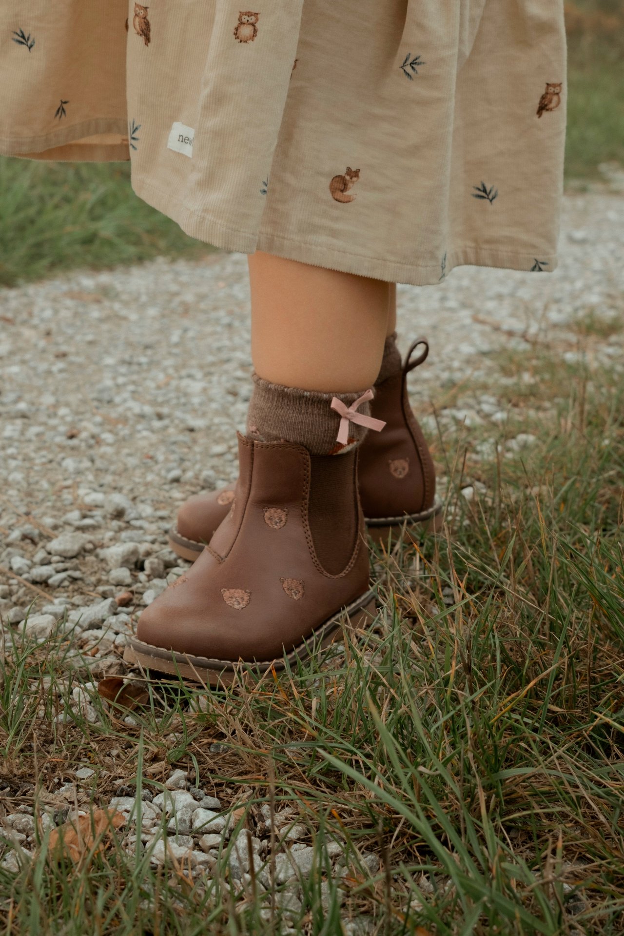 Nahaufnahme von Kinderstiefeln in Braun mit Teddybären, braunen Socken mit rosa Schleifen und beigem Cordkleid.