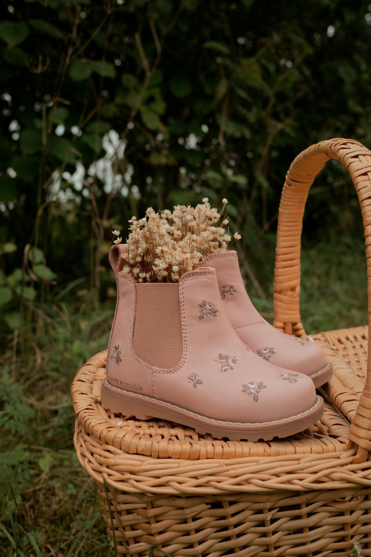 Nahaufnahme rosa Kinder-Chelsea-Stiefel mit Blumenstickerei und Trockenblumen, auf Weidenkorb.