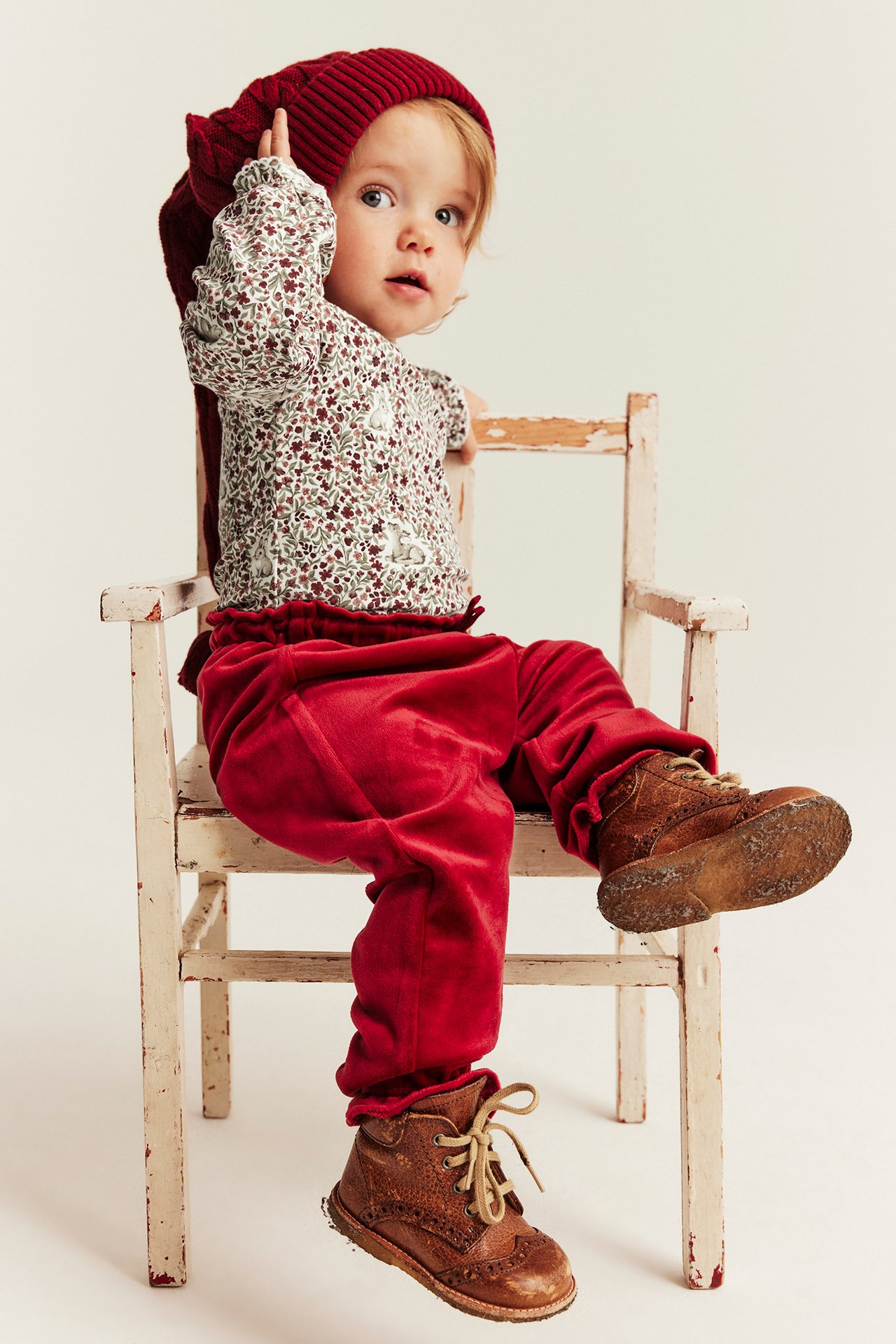 Baby in roter Strickmütze, weißem Blumen-Hasen-Oberteil, roter Samthose, braunen Stiefeln.