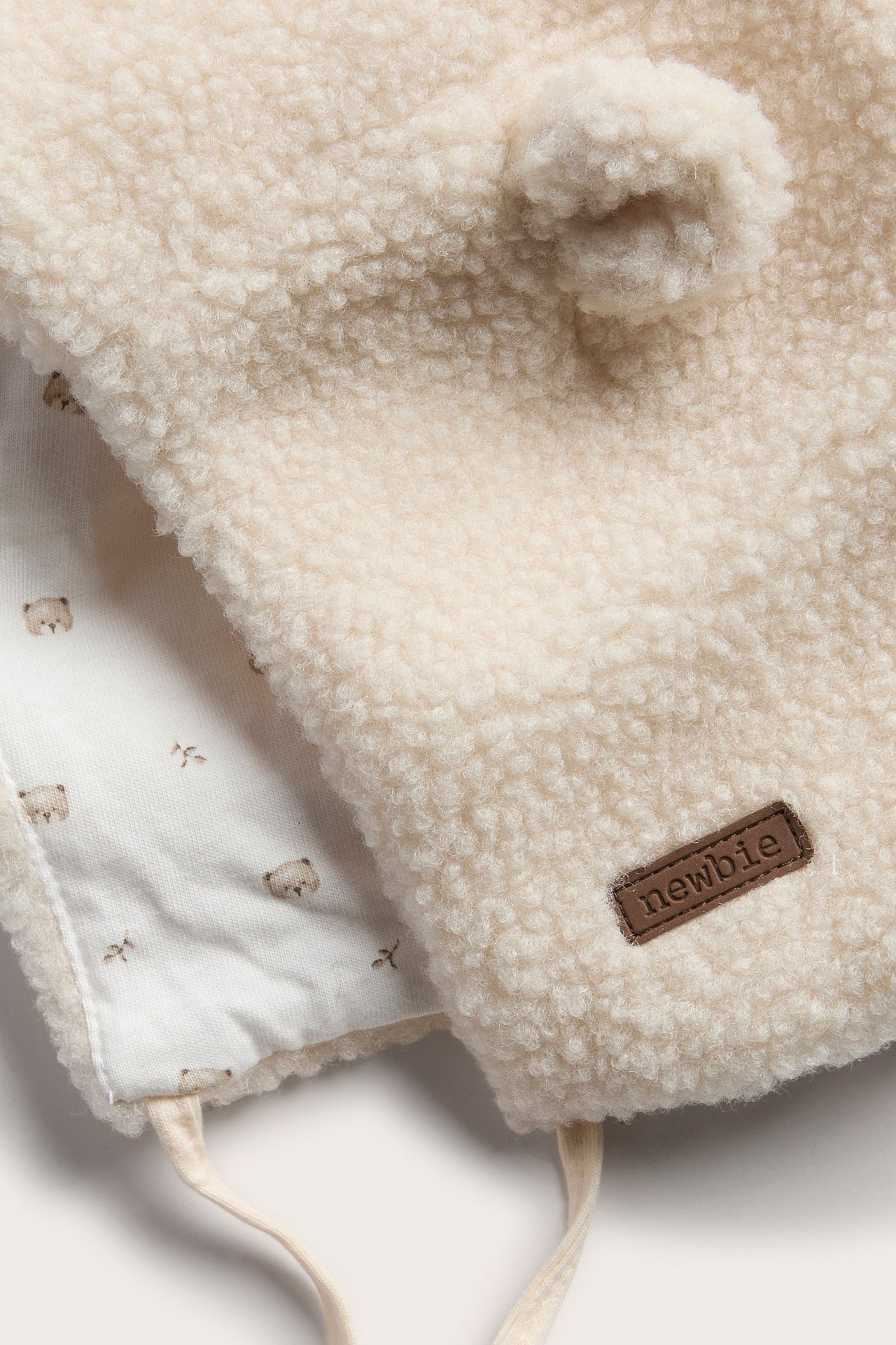 Nahaufnahme einer cremefarbenen Teddy-Fleece-Babyhaube mit braunem Newbie-Patch und gemustertem Futter.