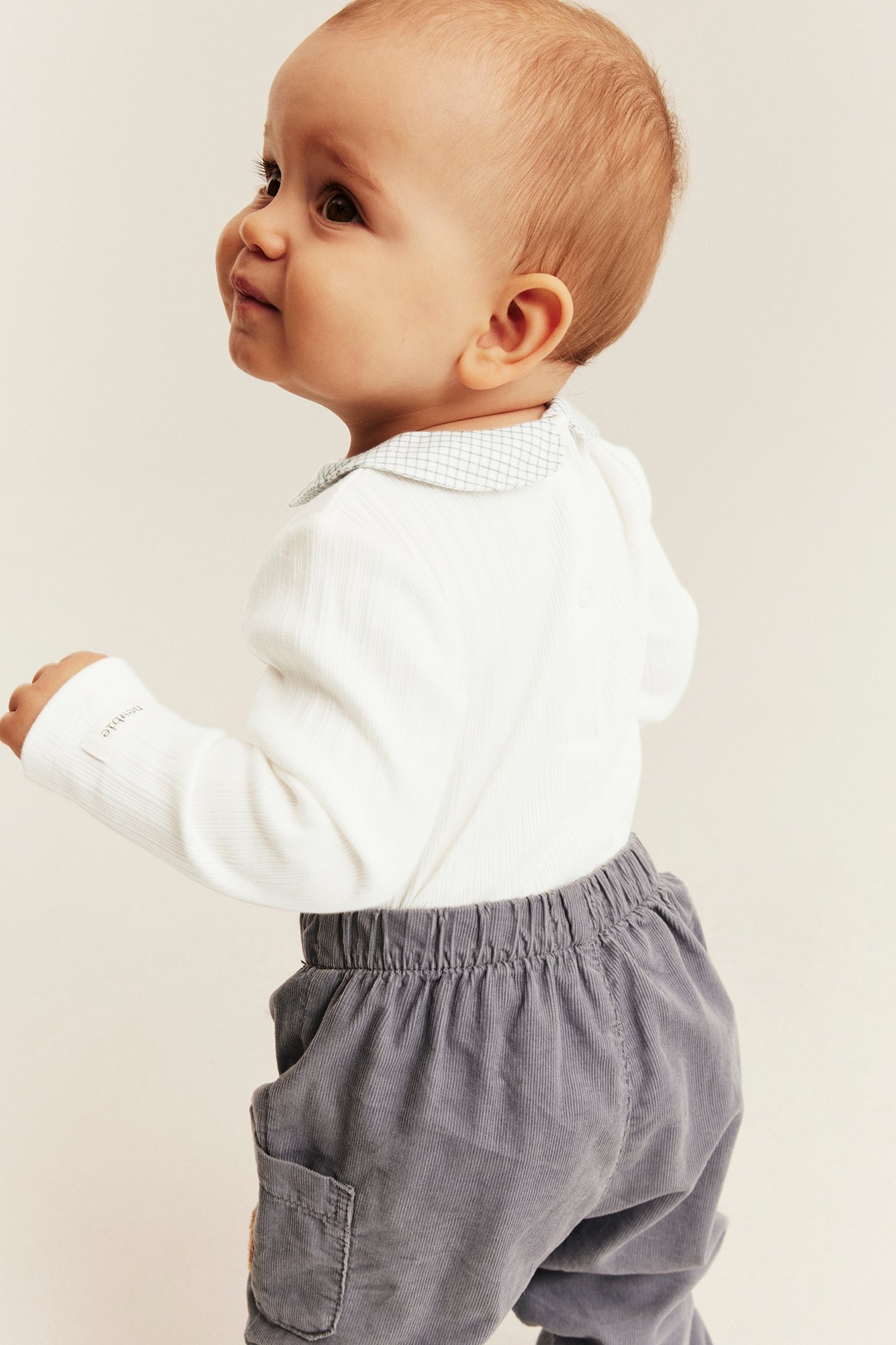 Back-side view of a baby wearing grey corduroy trousers and a white ribbed bodysuit with a patterned collar.