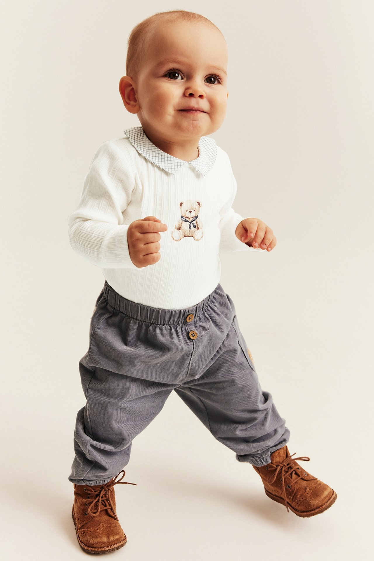 Front view of baby in a white ribbed teddy bear bodysuit, gray corduroy pants, and brown boots.
