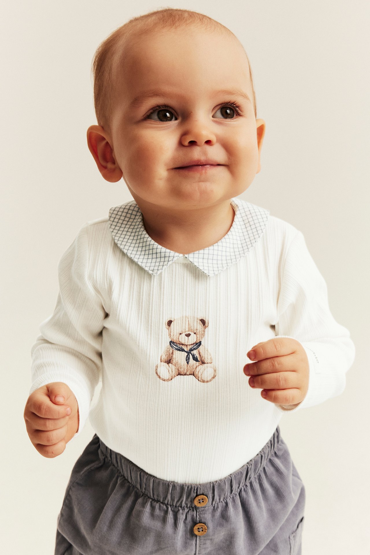 White ribbed long-sleeve bodysuit with teddy bear, plaid collar, grey corduroy pants on baby.
