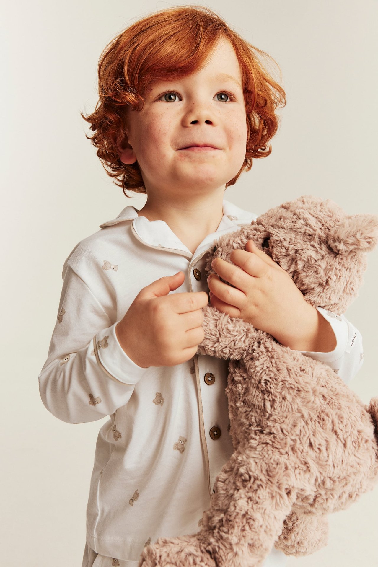 Kind in weißem langärmeligem Teddybär-Print Pyjama, hält einen braunen Plüsch-Teddybär.