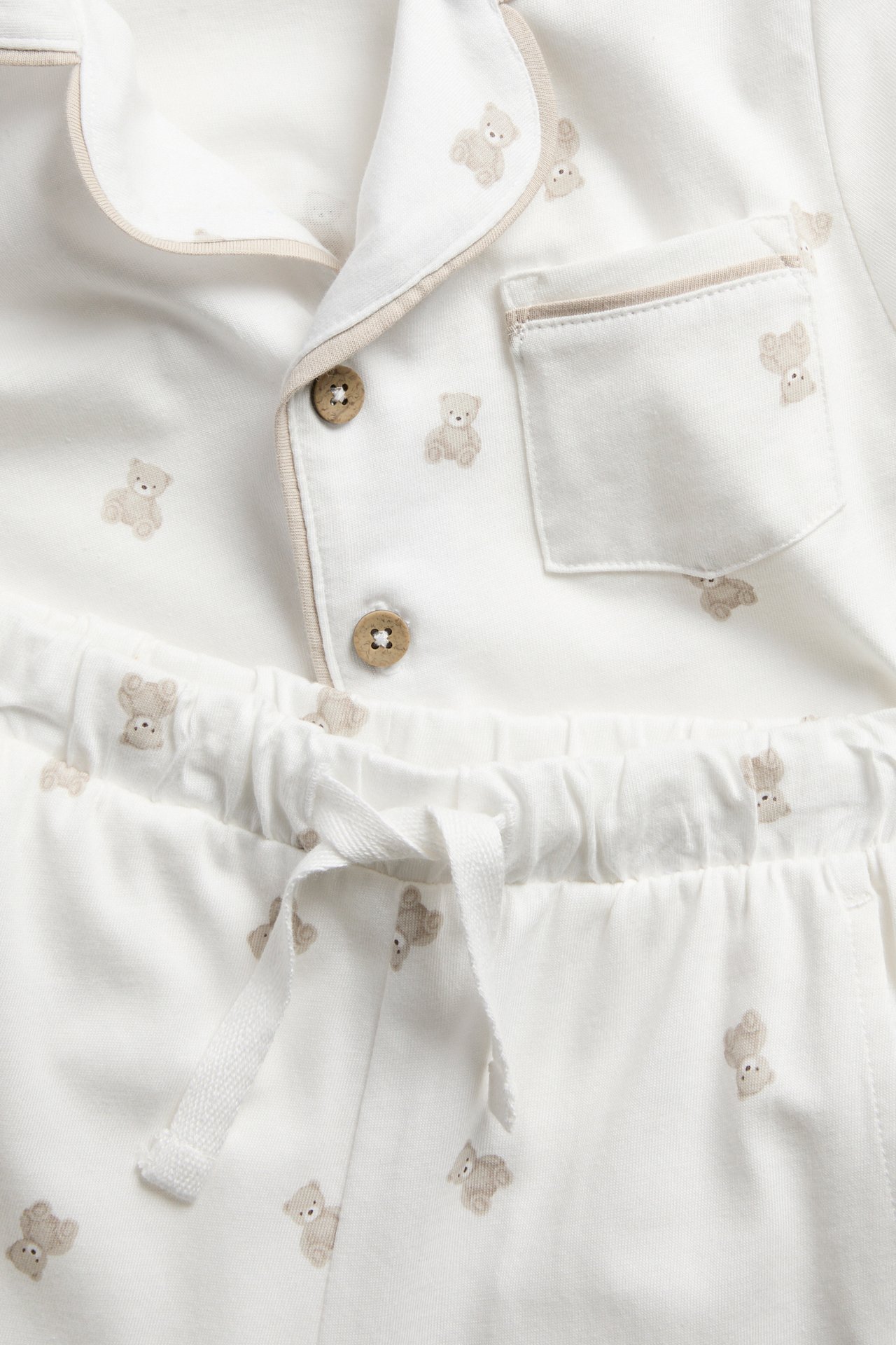 Nahaufnahme eines weißen Baby-Schlafanzug-Sets mit beigem Teddybär-Print und Knöpfen.