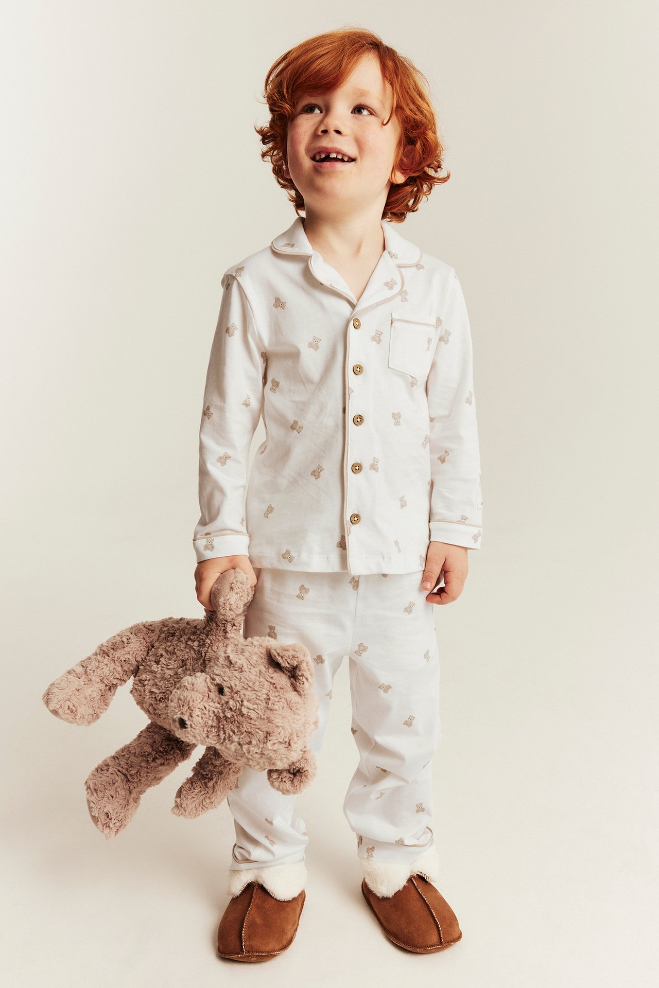 Vorderansicht Kind in weißem Teddybärmuster-Schlafanzug, hält braunen Teddy und braune Hausschuhe.