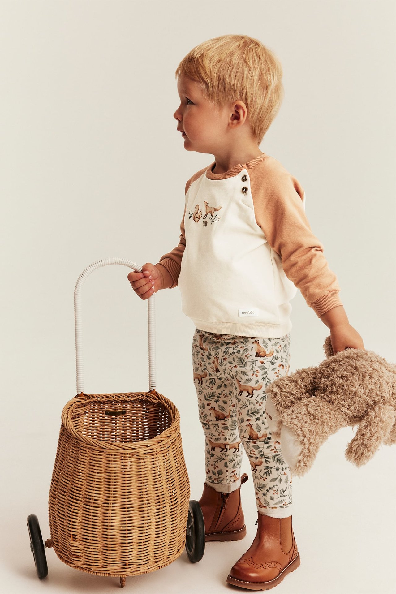 Side view of a kid in an embroidered sweatshirt, patterned leggings, brown boots, holding a wicker basket and toy.