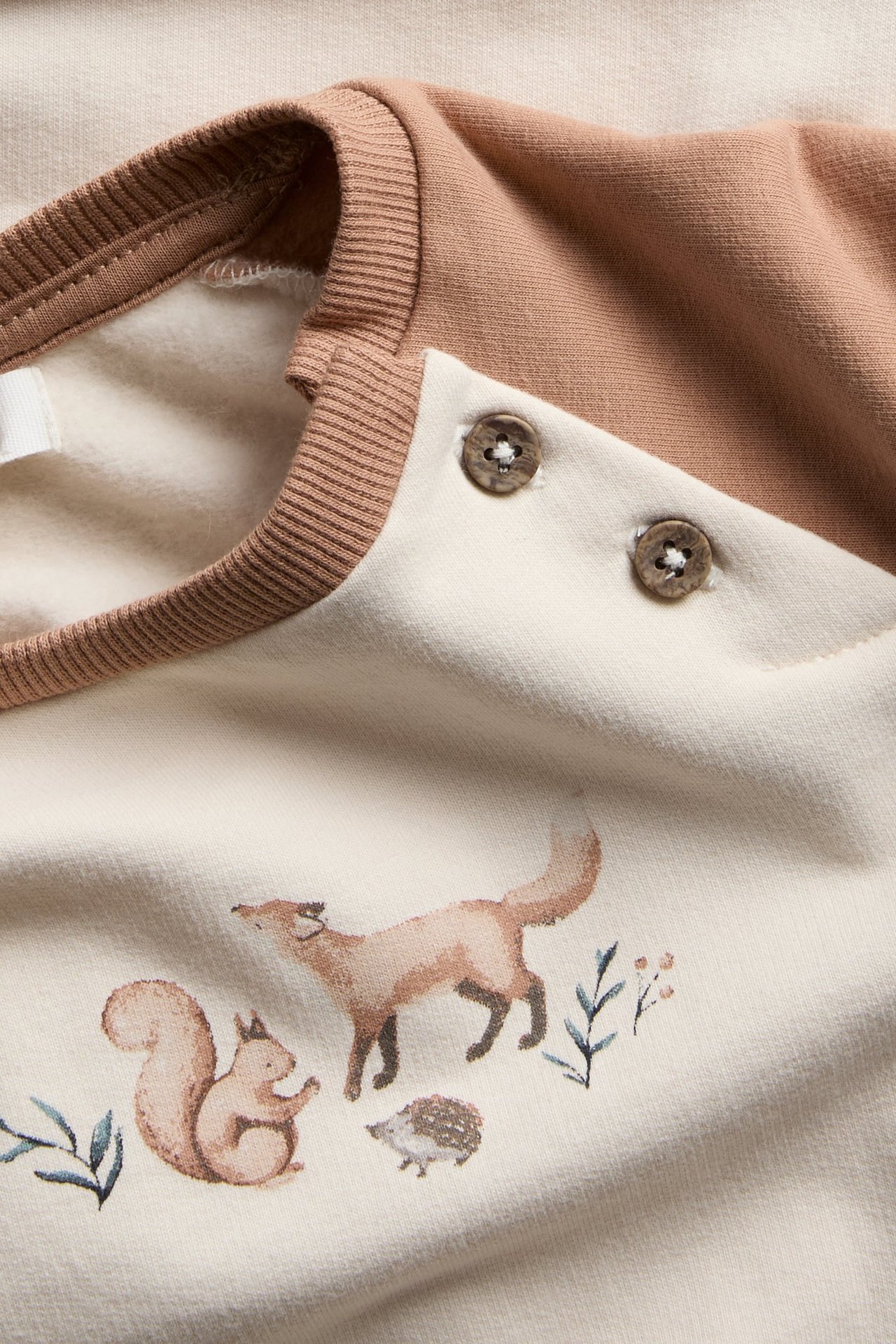 Nahaufnahme eines cremefarbenen Baby-Sweatshirts mit braunen Ärmeln, Schulterknöpfen und Waldtiermuster.