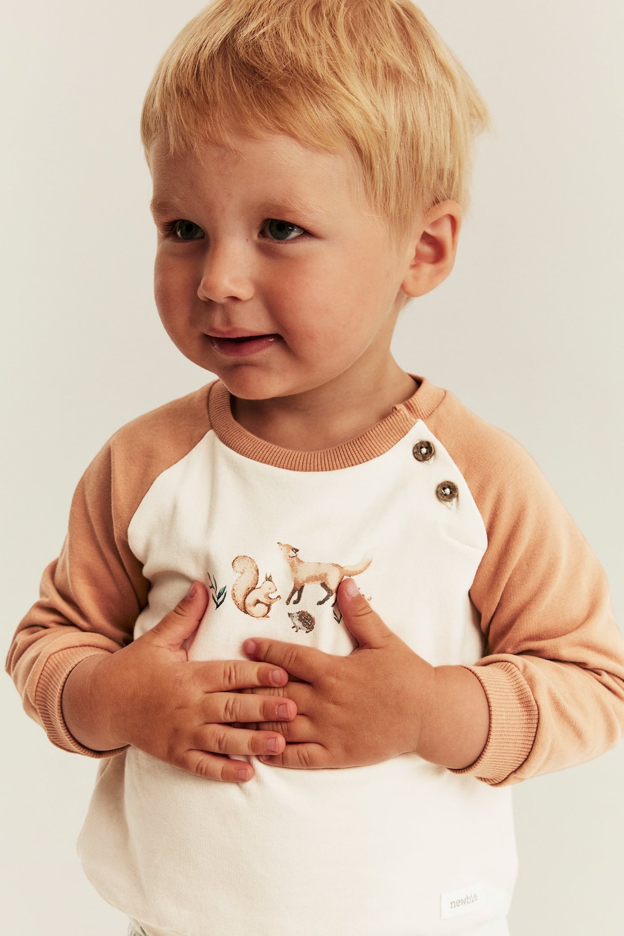 Front view of a kid wearing a cream and brown raglan sweatshirt with forest animal print and two buttons.