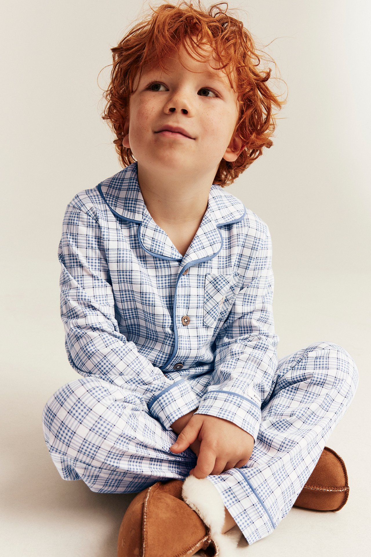 Kid in a blue and white plaid pajama set with buttons and brown sheepskin slippers, front view.