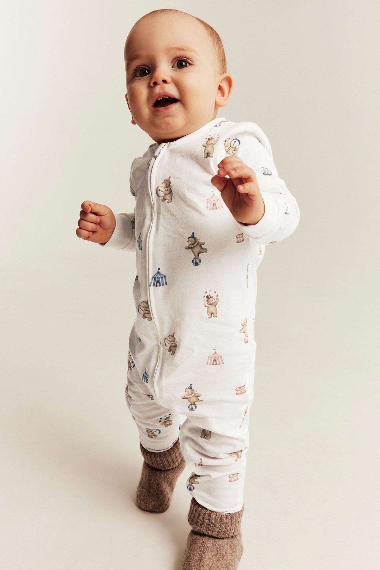 Vorderansicht eines Babys in weißem Overall mit Zirkustier-Print und braunen Strickschuhen.