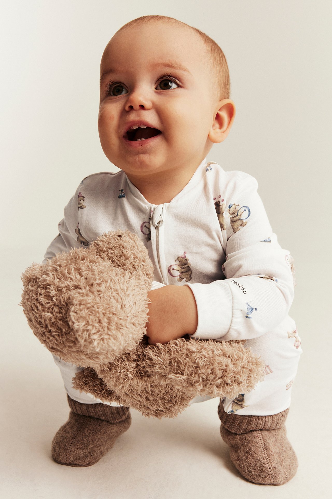 Vorderansicht eines Babys in weißem Overall mit Tiermuster, braunen Schühchen, hält einen braunen Teddy.