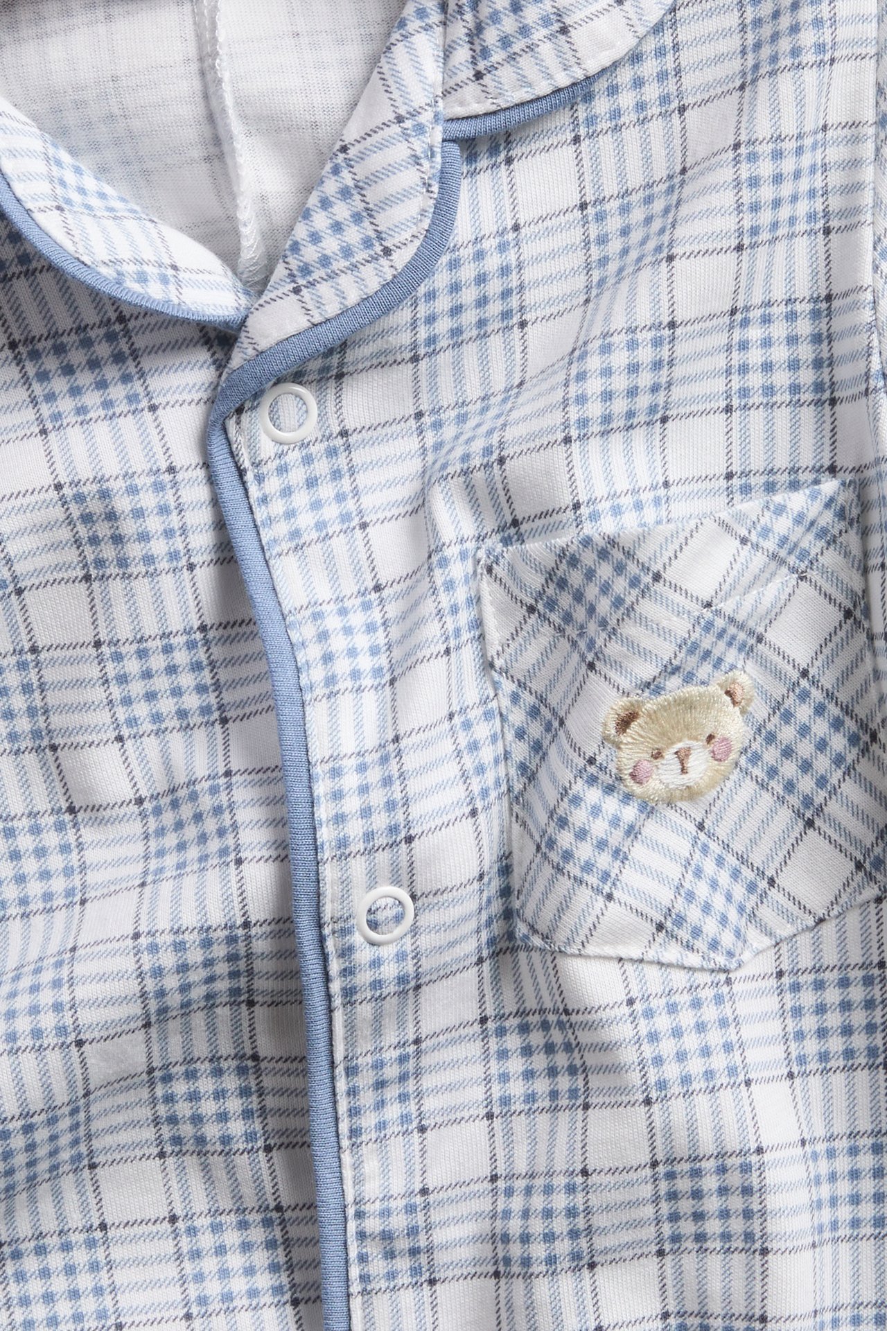 Nahaufnahme eines Baby-Schlafanzugoberteils in Weiß-Blau kariert mit blauer Paspelierung, Druckknöpfen und Teddy-Stickerei.