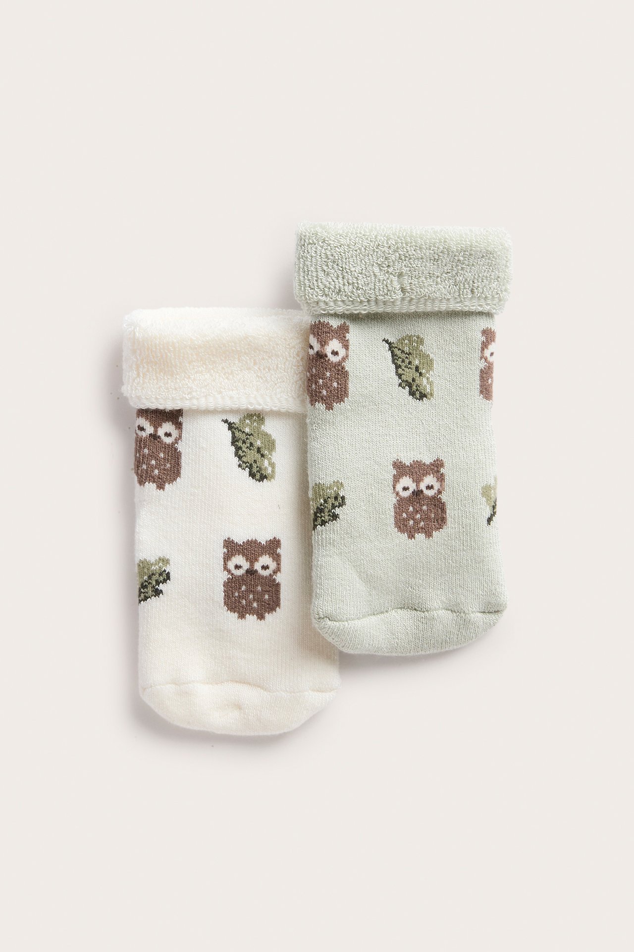 Draufsicht auf zwei Babysocken, cremeweiß und hellgrün, mit braunem Eulen- und grünem Blattmuster.