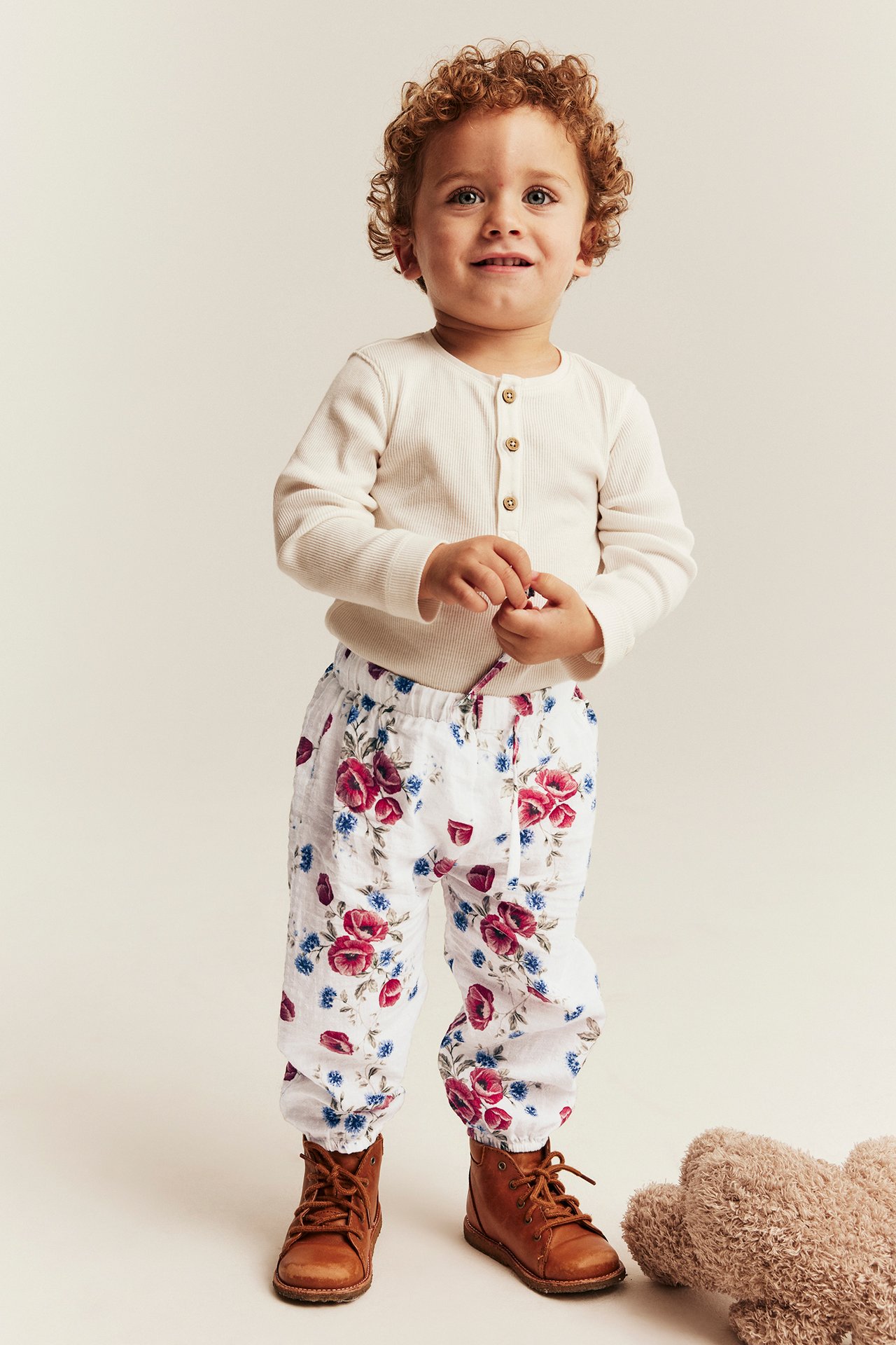 Front view of a baby wearing white floral print pants, a cream Henley shirt, and brown boots.