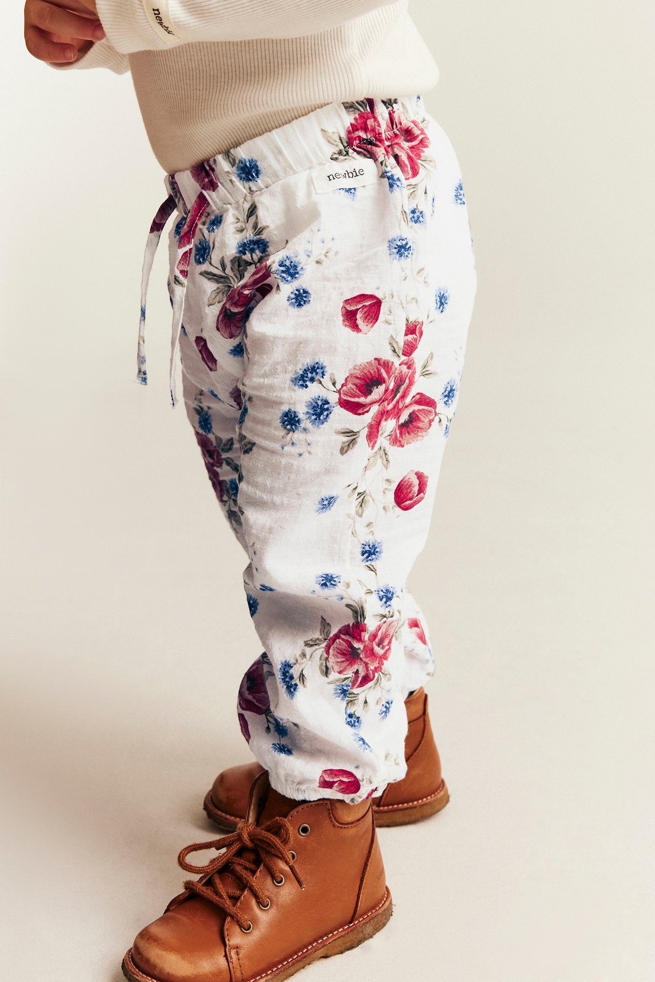 Seitenansicht von Baby in weißen Blumenmusterhosen, cremefarbenem Ripp-Top und braunen Schnürstiefeln.