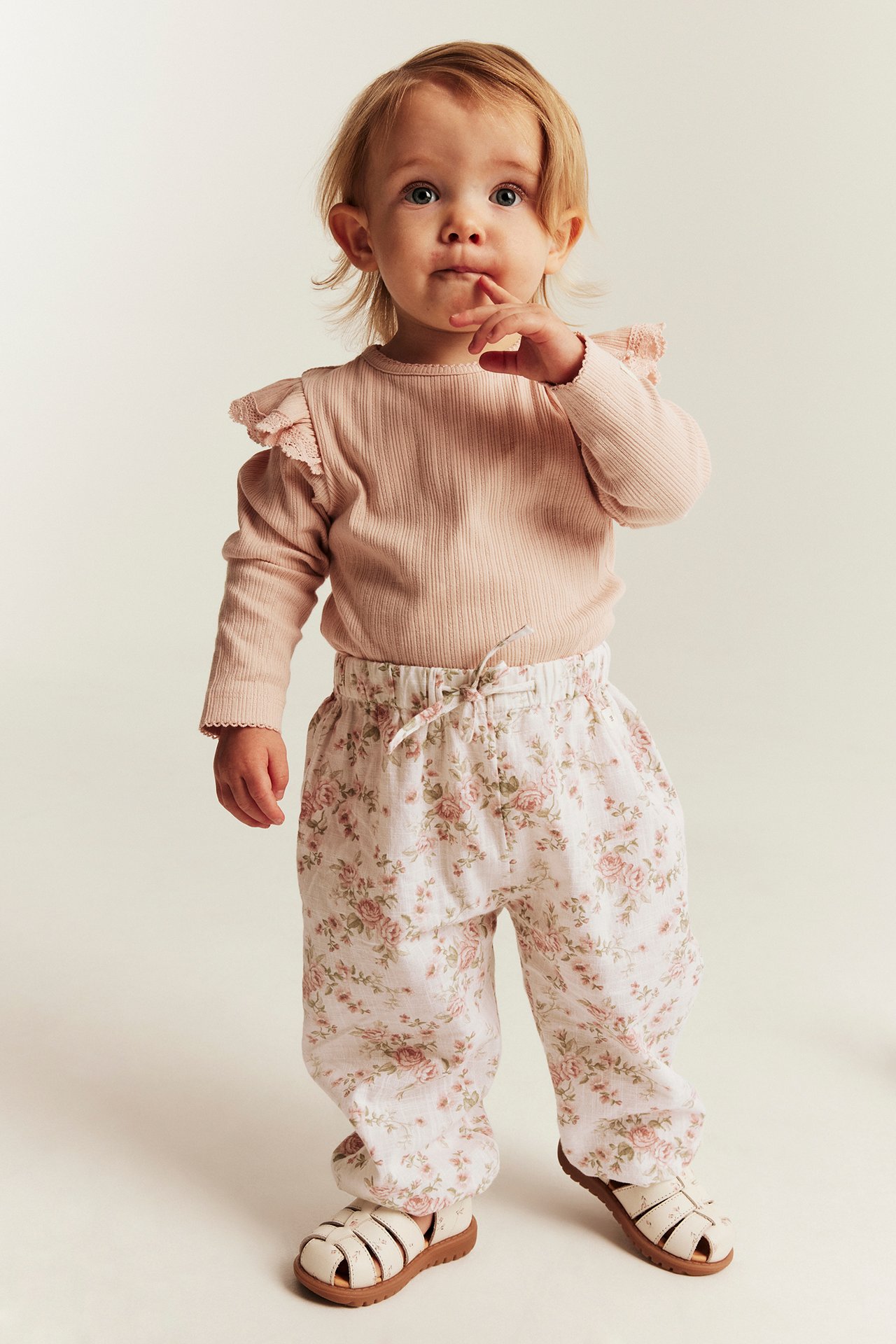Vorderansicht eines Babys in hellrosa geripptem Rüschen-Top, geblümter Hose und weißen Sandalen.