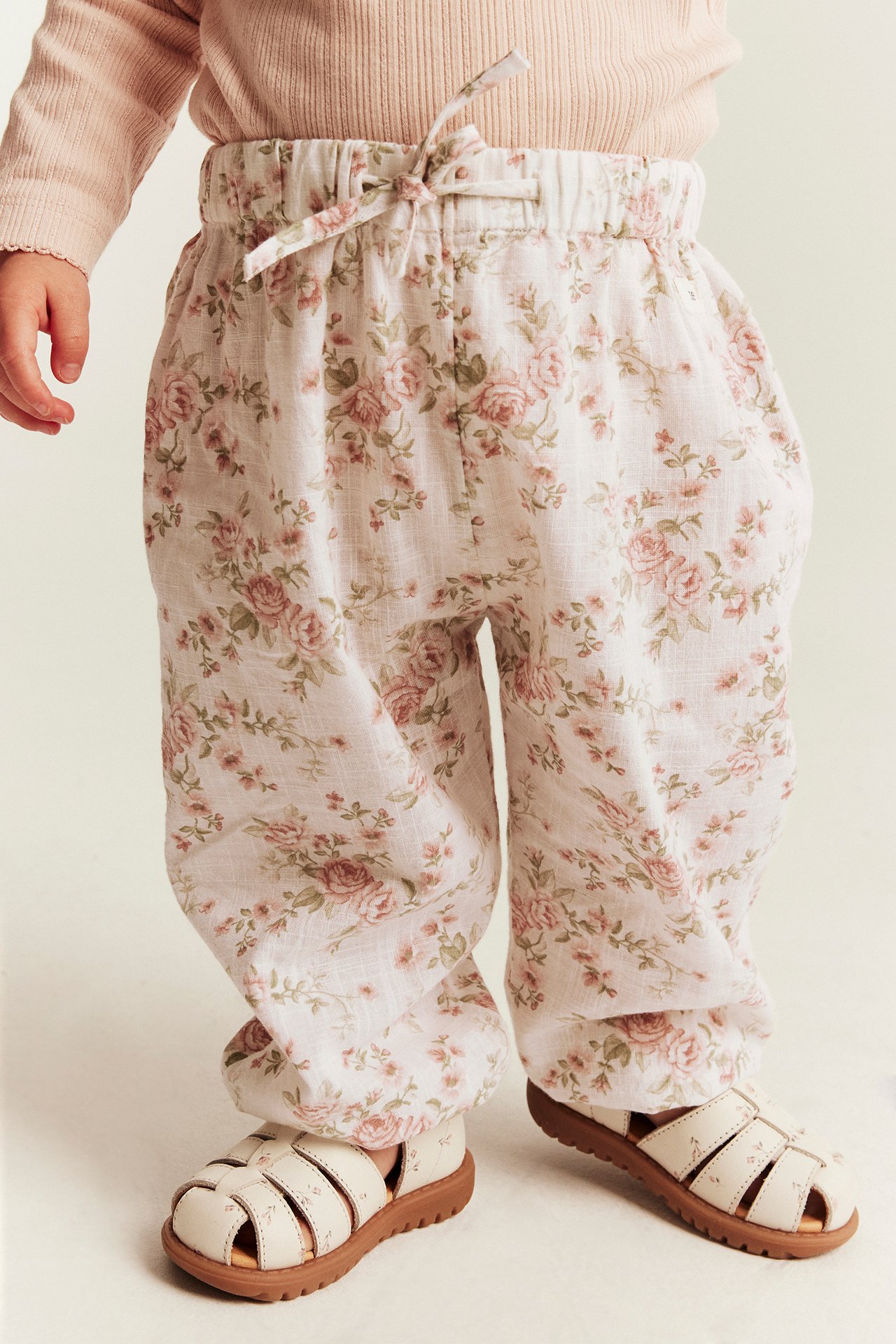 Vorderansicht von Baby in rosa geblümter Hose mit Schleife und cremefarbenen Sandalen.
