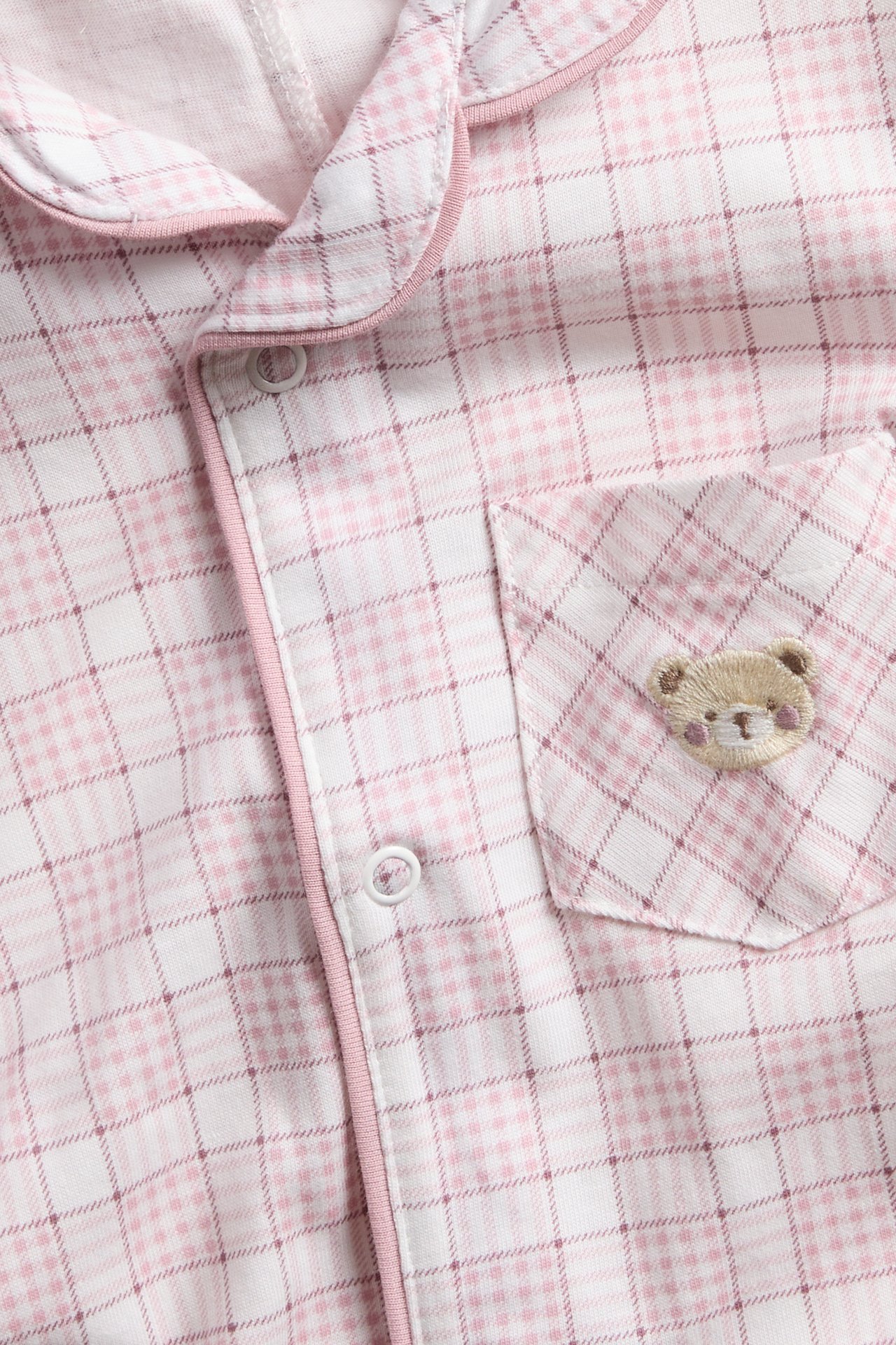 Close-up: Pink and white plaid pajama top with teddy bear embroidery on pocket, white buttons.