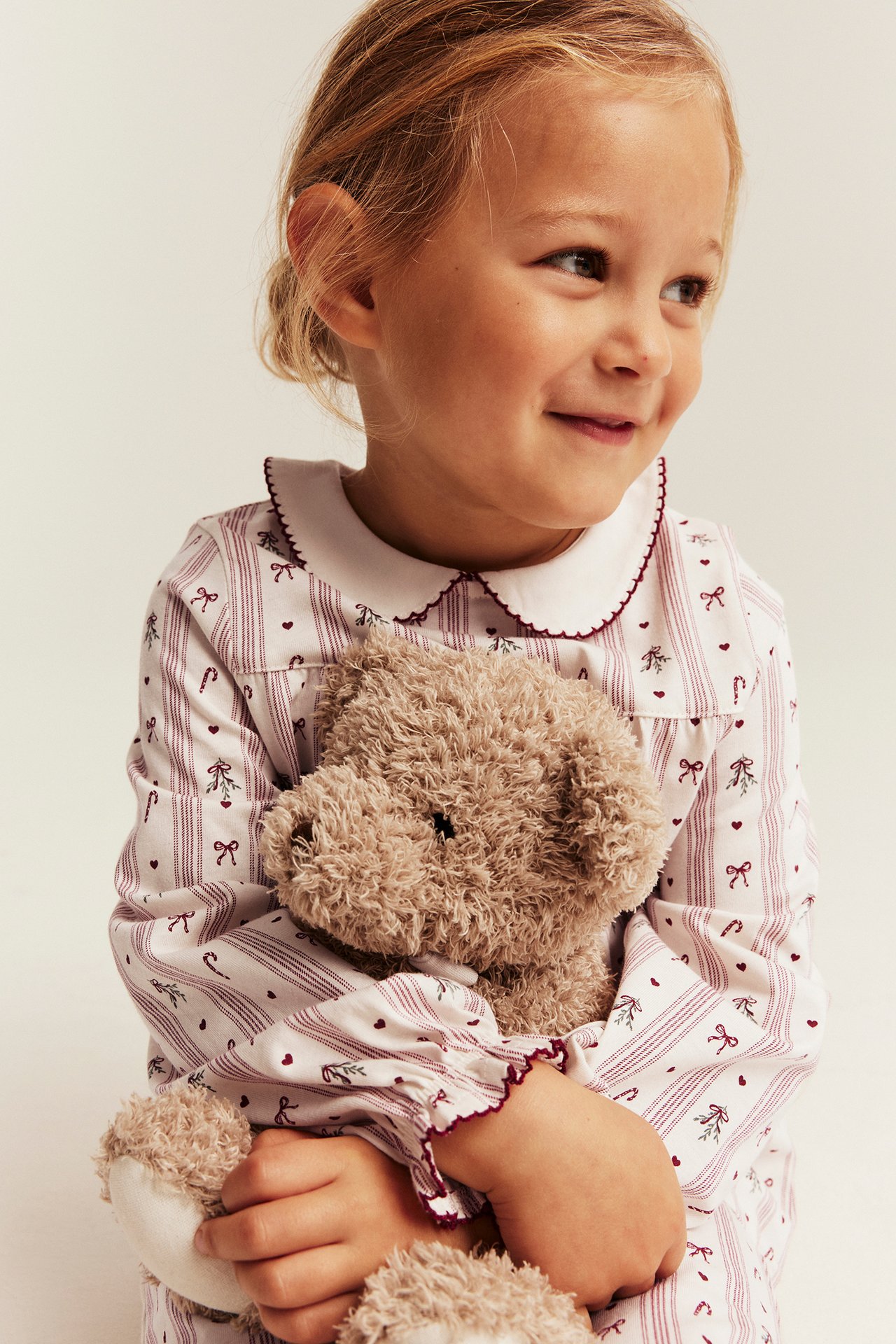 Kinder-Schlafanzug, weiß mit roten Streifen und Weihnachtsmotiven, rot eingefasst, hält braunen Teddybär.