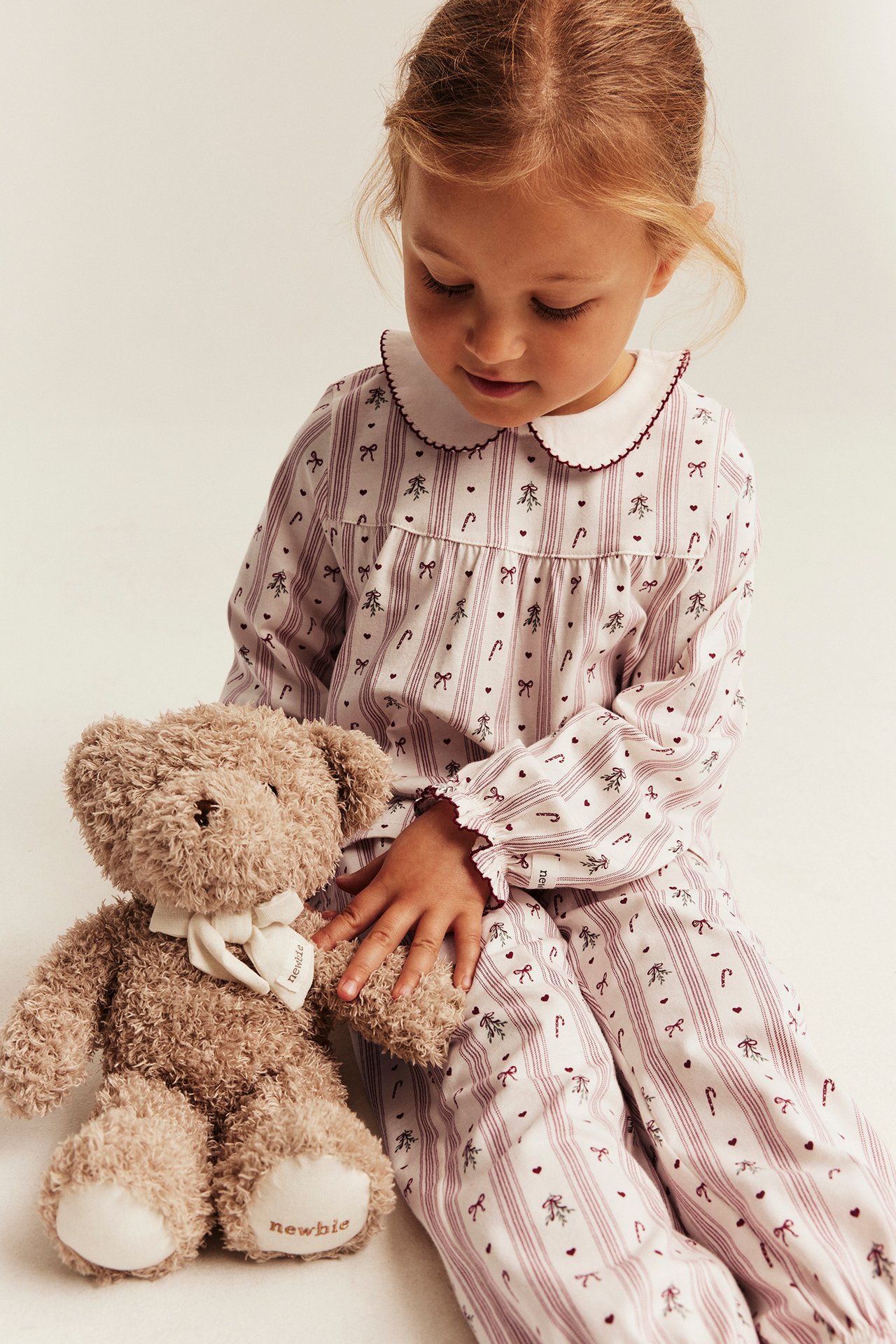 Vorderansicht, Kind in weißem Pyjama mit roten Streifen, Schleifen, Zuckerstangen, hält braunen Teddybär.