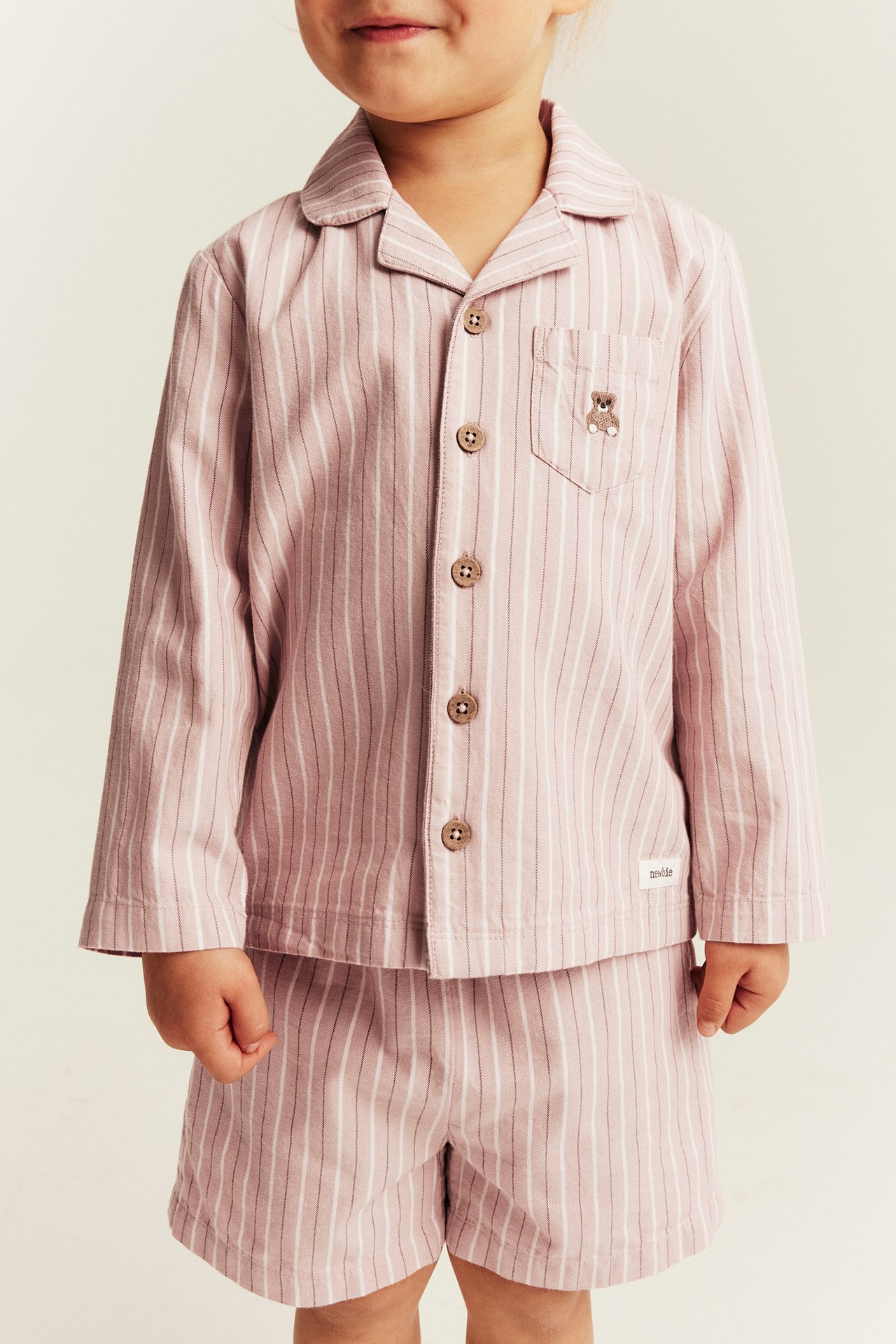 Front view of a kid wearing a pink and white striped pajama shirt with shorts, featuring buttons and a bear embroidery.