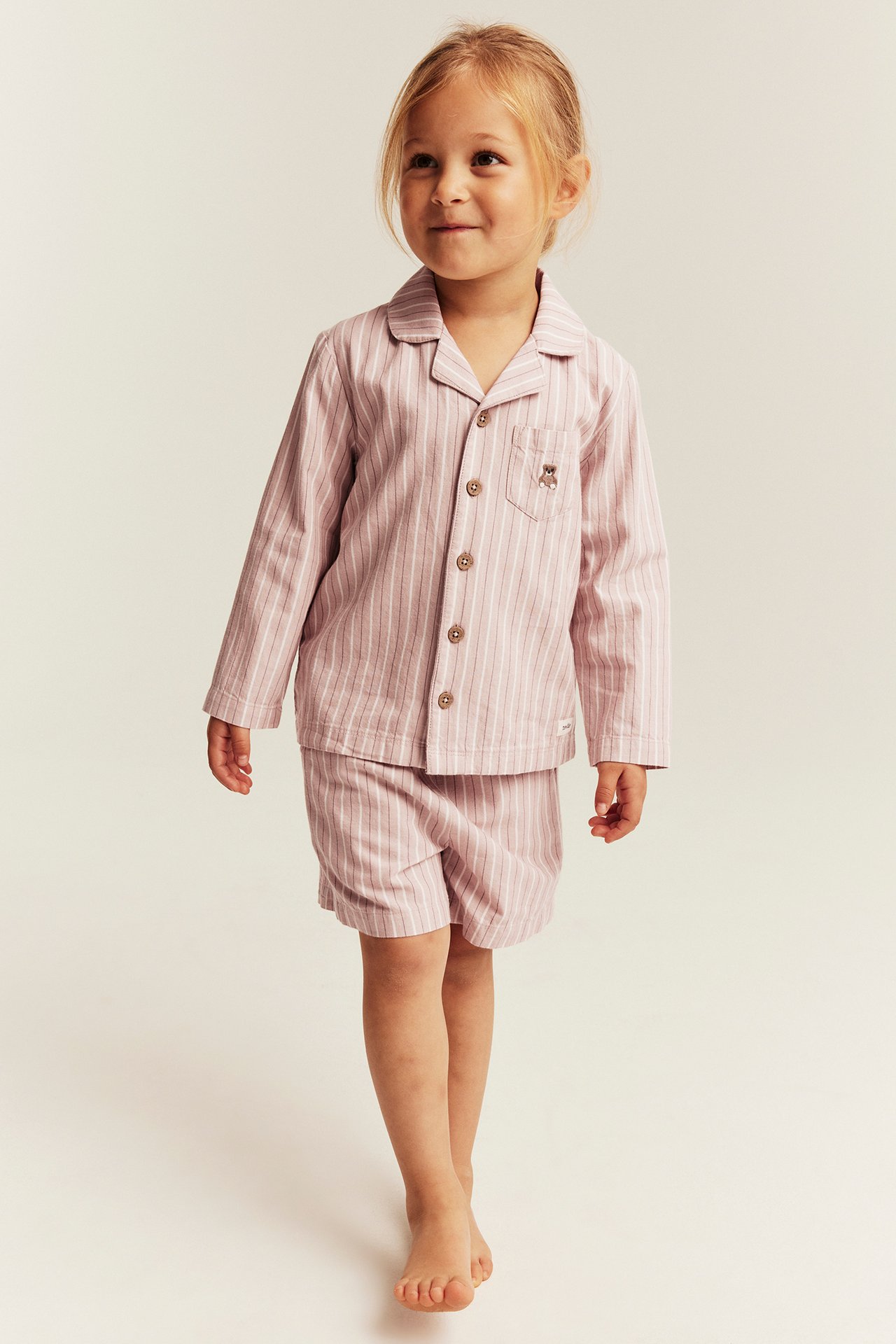 Vorderansicht: Kind in rosa gestreiftem Pyjama-Set, Langarmshirt mit Bärenstickerei und Shorts.