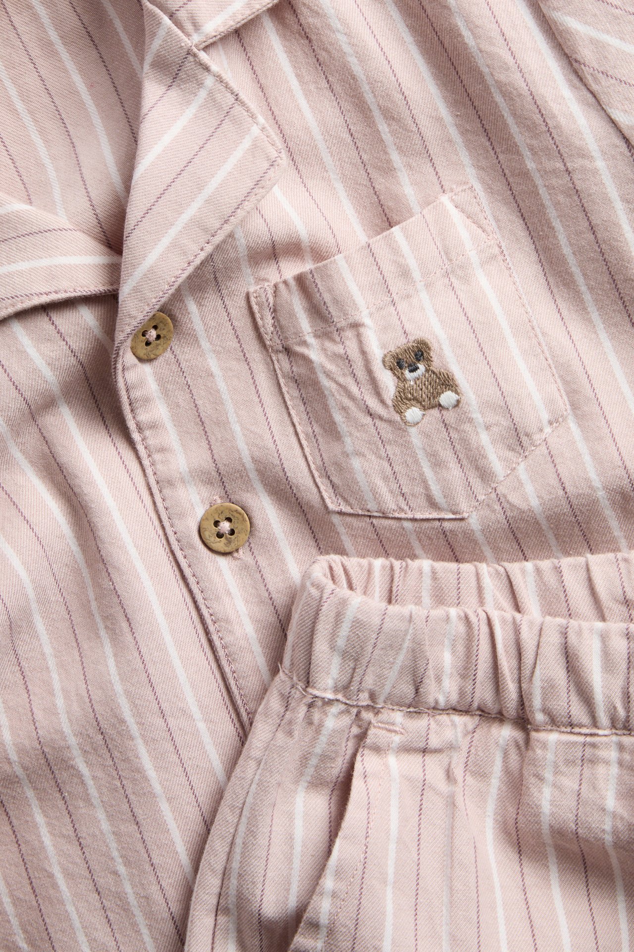 Nahaufnahme eines rosa gestreiften Pyjama-Sets mit Knöpfen und einem gestickten Teddybär auf der Tasche.