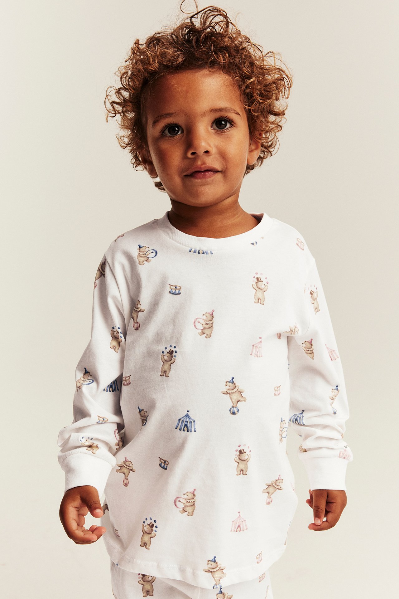 Vorderansicht eines Kindes in weißem Langarmshirt mit Zirkusbären-Print und weißen Hosen.