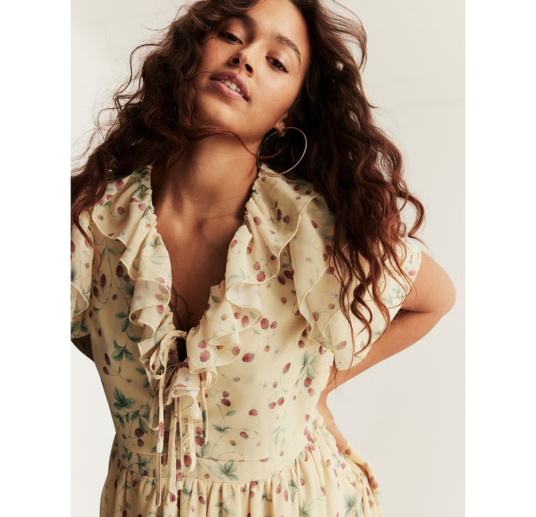 Woman wearing a light floral dress with ruffled neckline, tie detail at the front, and soft, flowing fabric.