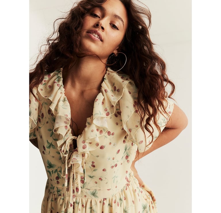 Woman wearing a light floral dress with ruffled neckline, tie detail at the front, and soft, flowing fabric.